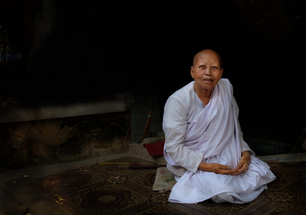 A female Cambodian Buddhist nun wears white robes and kneels on the floor with her hands folded on her lap.